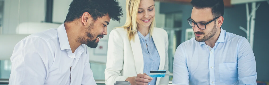 Three colleagues in a modern office smiling and looking at a credit card held by one of them.