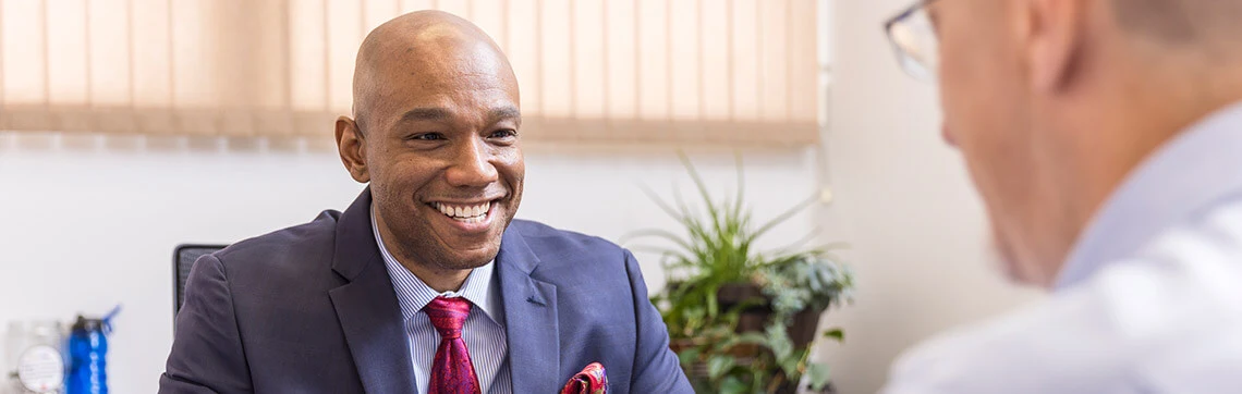 Smiling man in a suit talking with another person in an office setting.