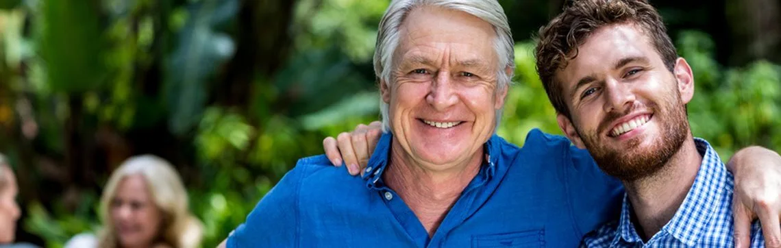 A smiling older man in a blue shirt and a younger man in a checkered shirt pose happily together outdoors, with lush greenery in the background.