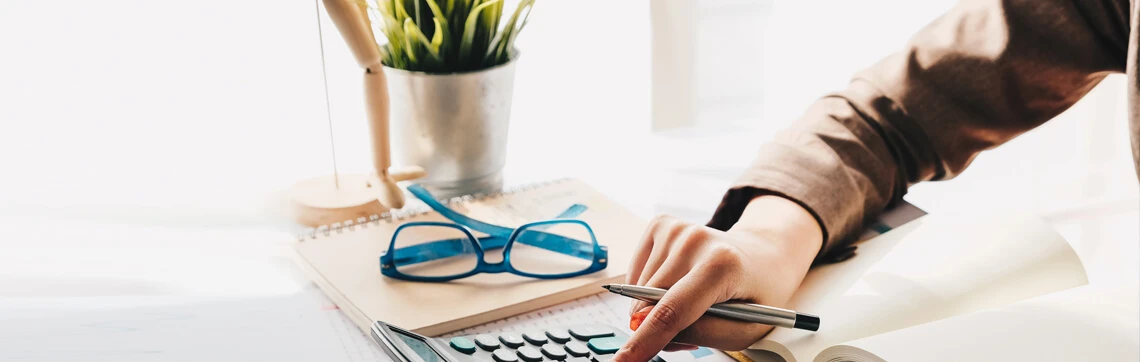 A hand with a pen points at a calculator, surrounded by a notebook, blue glasses, and a potted plant. The setting is bright, conveying focus.