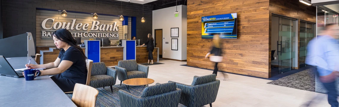 Modern bank lobby with a sleek design. A woman works on a laptop at a communal table. Customers approach the counter. Blue and wood tones create a welcoming atmosphere.
