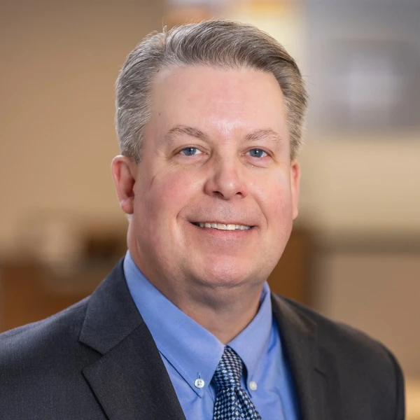 Man smiles in front of a blurred background. He is wearing a suit and tie.