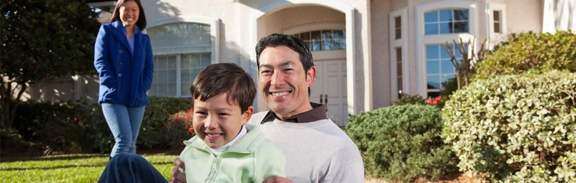 Smiling man and child sitting on lawn with woman standing behind them in front of a house.