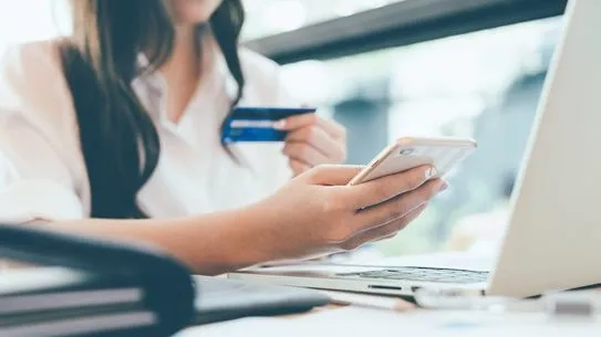 A woman using a smartphone holds a credit card beside a laptop on a desk, suggesting online shopping. The scene appears focused and modern.