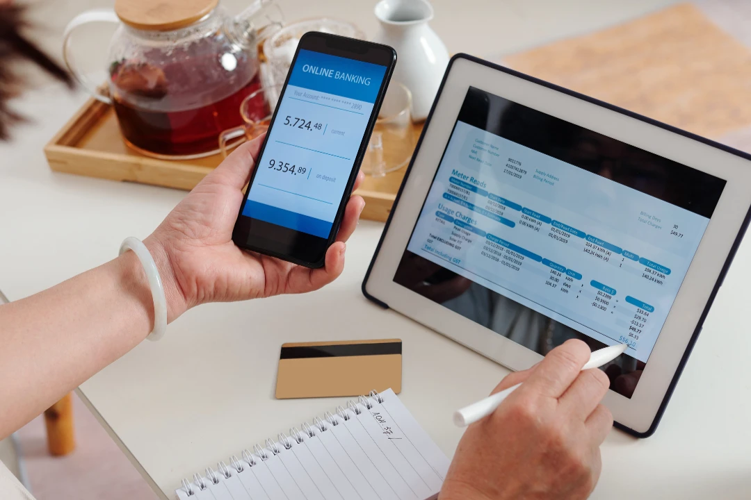 Person managing finances with a smartphone displaying online banking, a tablet showing a utility bill, and a notepad on a table with a credit card nearby.