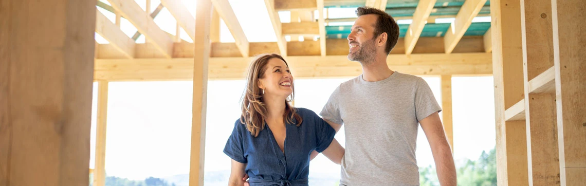 Couple smiling and looking around inside a wooden house under construction.