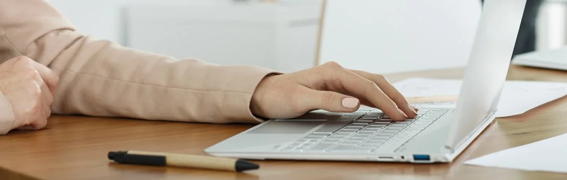 Close-up of a person’s hand typing on a laptop at a wooden desk with a pen nearby.