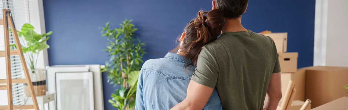 Couple stands in front of a blue wall with house plants and moving boxes scattered around