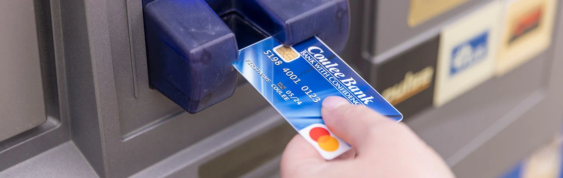 A hand inserts a credit card into an ATM. The card is blue with banking details visible. The image conveys a focused, transactional tone.