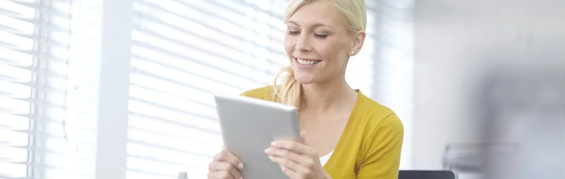 Smiling woman in a bright office using a digital tablet near a window with blinds.