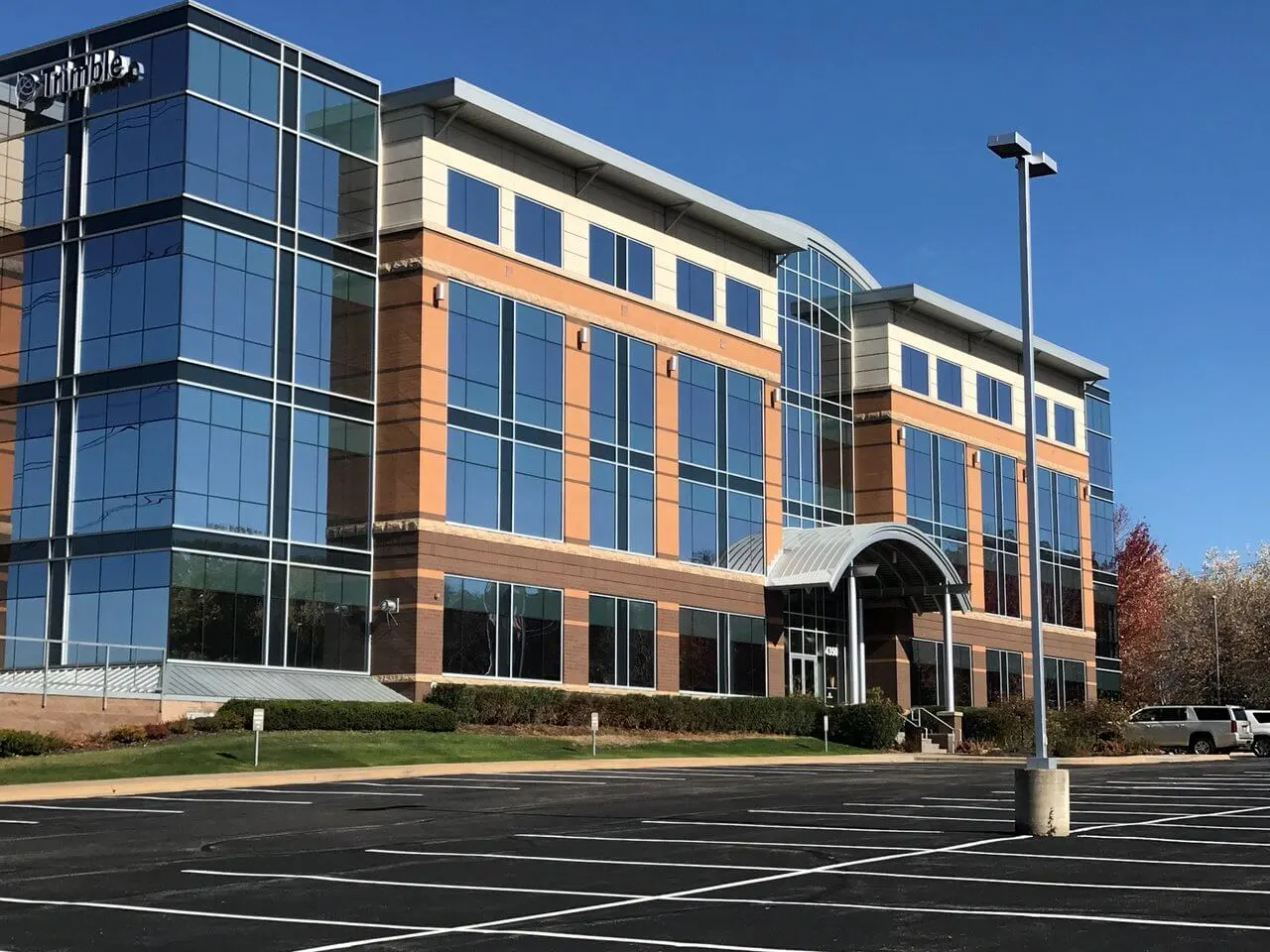 A modern, four-story office building with large glass windows and brick facade reflects the clear blue sky. Empty parking lot and light poles in front.