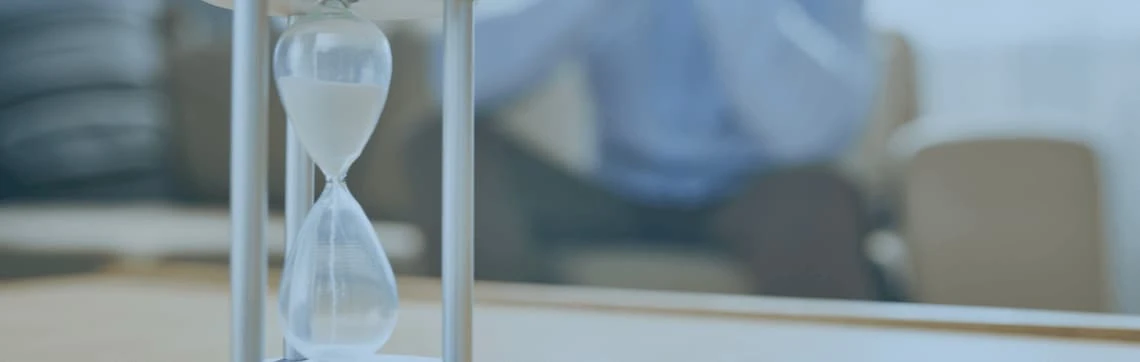 A close-up of a sand-filled hourglass on a table, with sand trickling down. A blurred figure in the background conveys a contemplative mood.