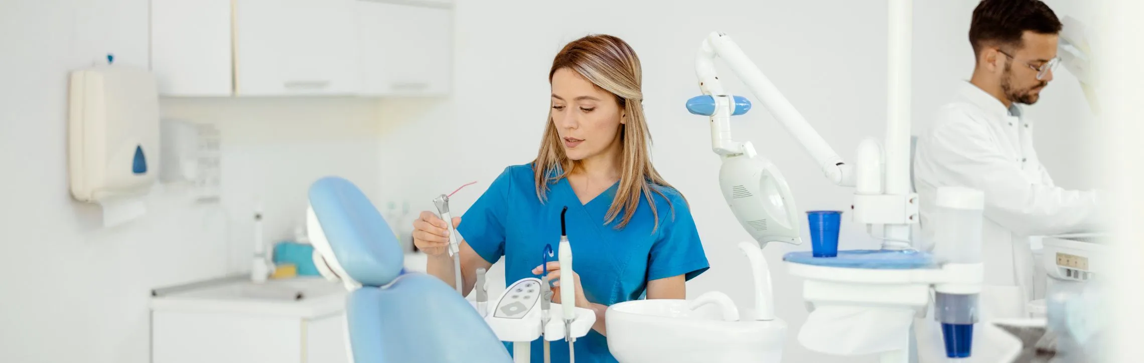 Dentist is seen organizing tools next to a dental chair. There is another dentist in the background.