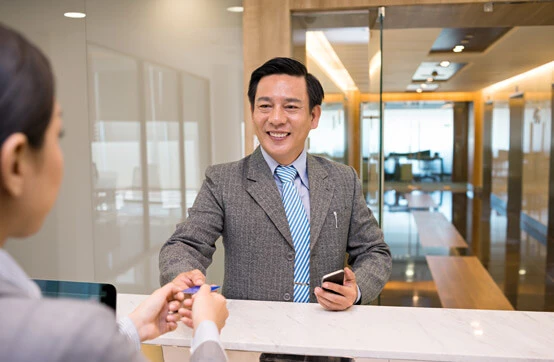 Smiling businessman at a reception desk handing over a pen, holding a phone in his other hand.