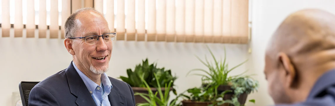 Smiling man in a suit talking to another person in an office with plants in the background.