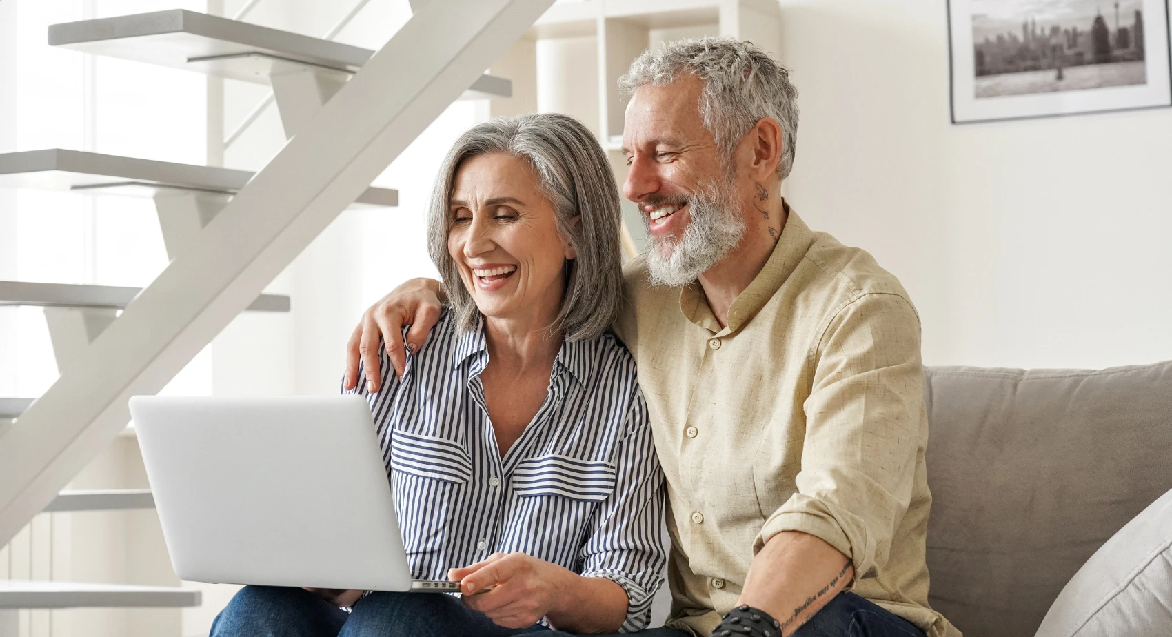 A happy older couple sits on stairs, smiling and looking at a laptop. The background is a bright, airy home.