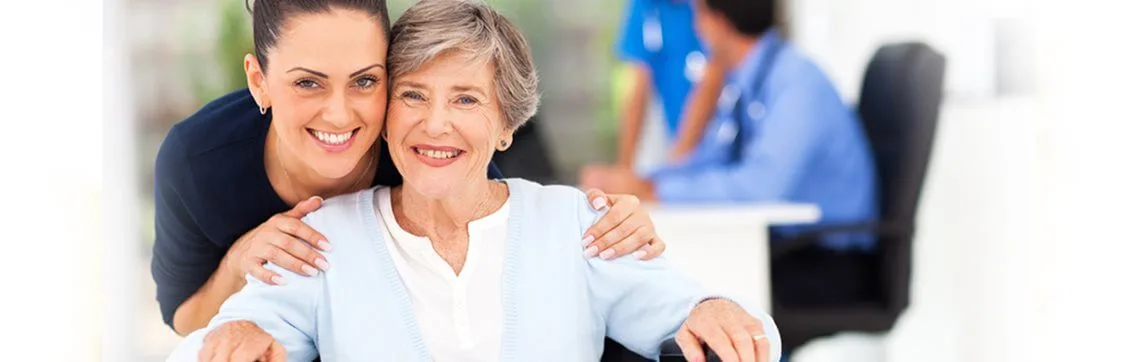 A smiling caregiver embraces an elderly woman in a light blue sweater. A doctor in a blue coat is seen in the blurred background. Warm, supportive atmosphere.