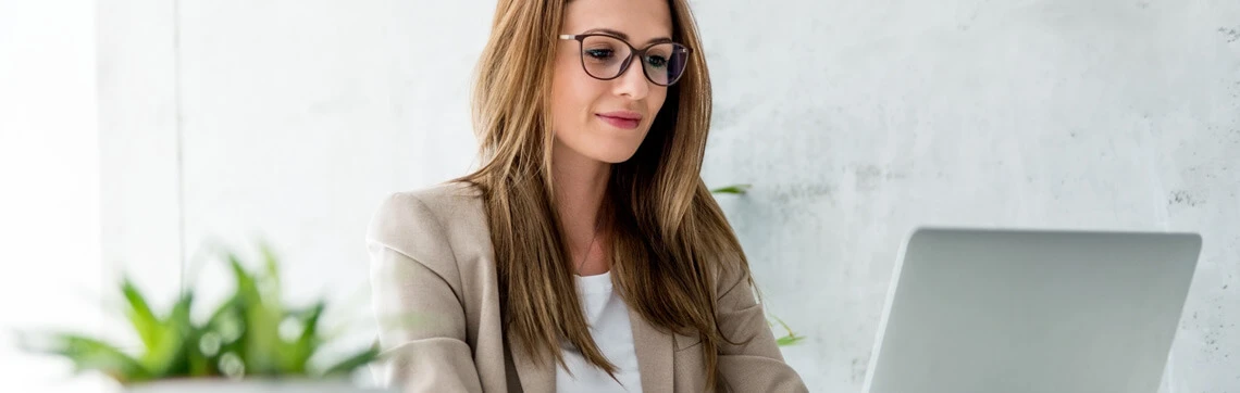 A professional woman with long brown hair and glasses is sitting at a desk, working on a laptop.