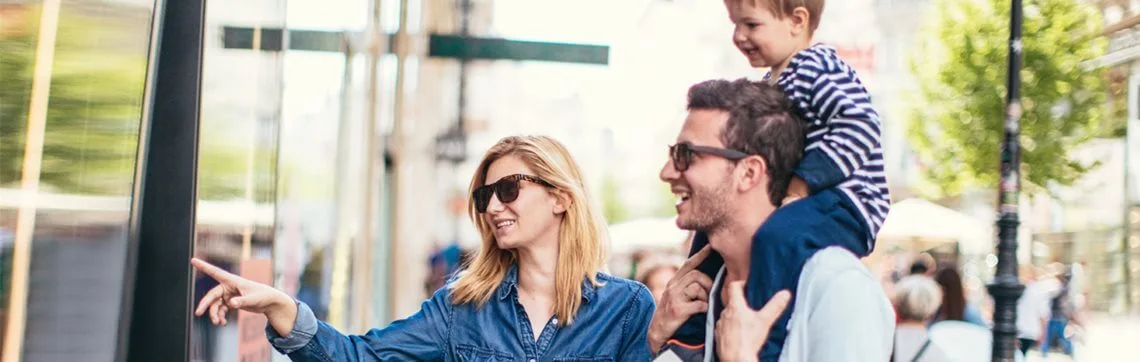 A smiling family enjoys a sunny day outside, with a woman pointing, a man carrying a child on his shoulders. All wear sunglasses, capturing a joyful moment.