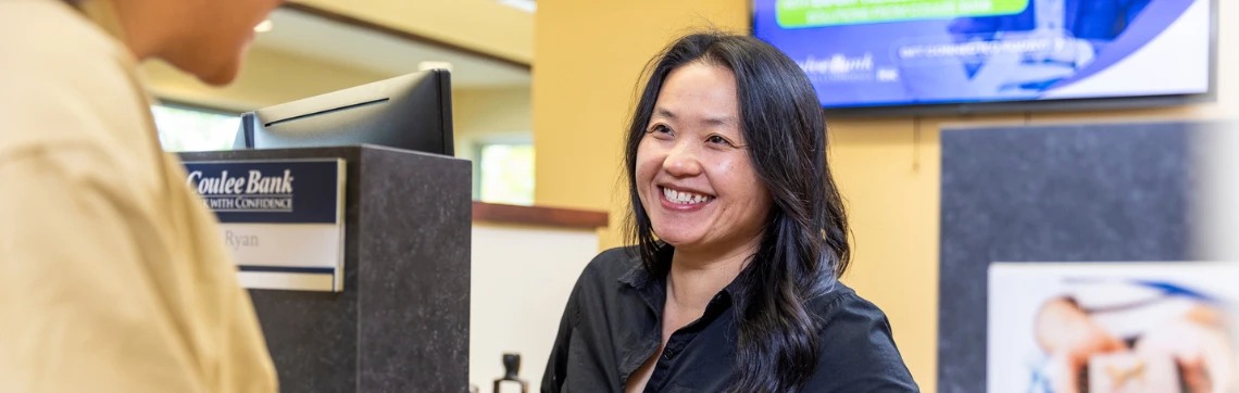 A smiling bank teller with long dark hair, wearing a black shirt, assists a customer at the counter. The atmosphere is welcoming and professional.