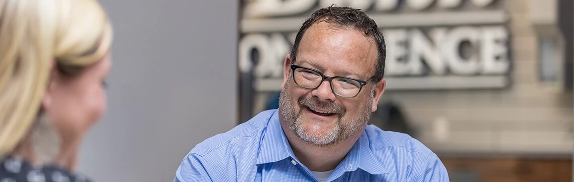 A man with glasses and a blue shirt smiles warmly during a conversation. The background shows a blurred sign, conveying a friendly, professional atmosphere.