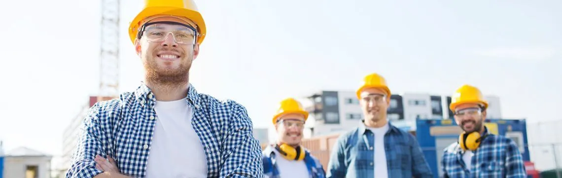 Group of construction workers wearing hard hats and safety gear at a construction site, with one worker smiling in the foreground.