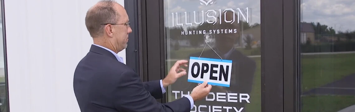 A man hangs an "OPEN" sign on the glass door of a business named "Illusion Hunting Systems – The Deer Society."
