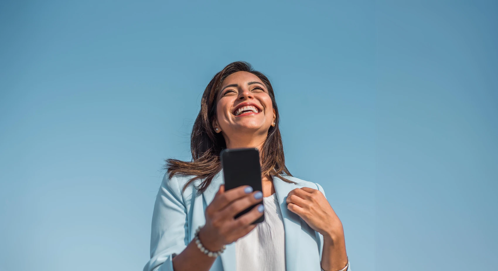 Smiling woman in a light blue blazer holding a smartphone, standing against a clear blue sky. She exudes happiness and confidence.