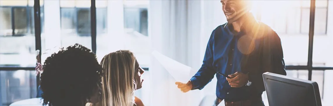 Man smiling and handing a document to two colleagues during a meeting in a brightly lit office.
