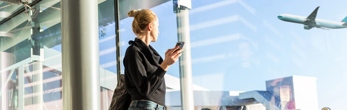 A woman in a black sweater holding a smartphone gazes out a large airport window. A plane is visible in the sky, conveying a sense of travel and anticipation.
