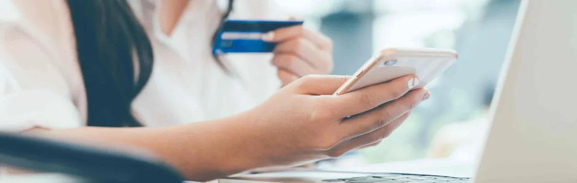 A person holding a smartphone and a credit card while sitting at a laptop, suggesting online banking or shopping.