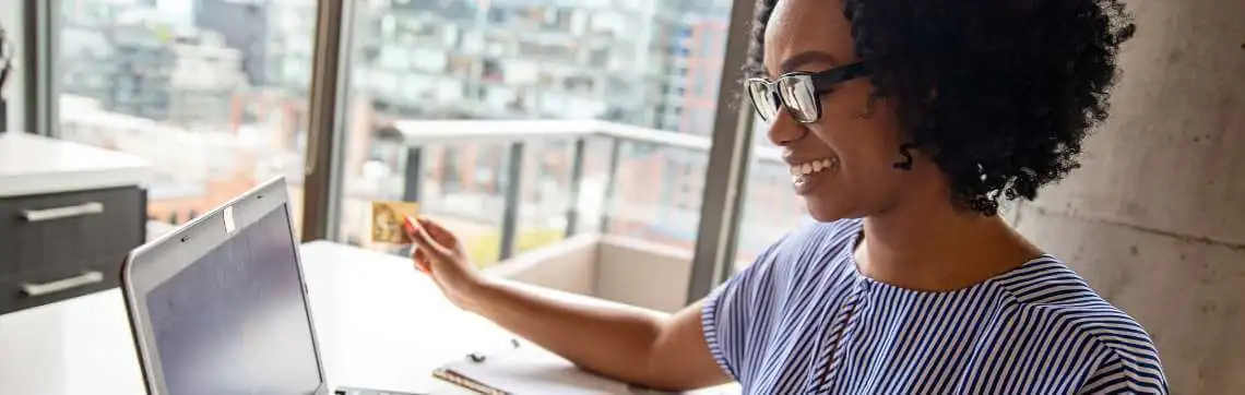 A woman with curly hair and glasses, smiling as she holds a credit card, works on a laptop in a bright office with a cityscape view.