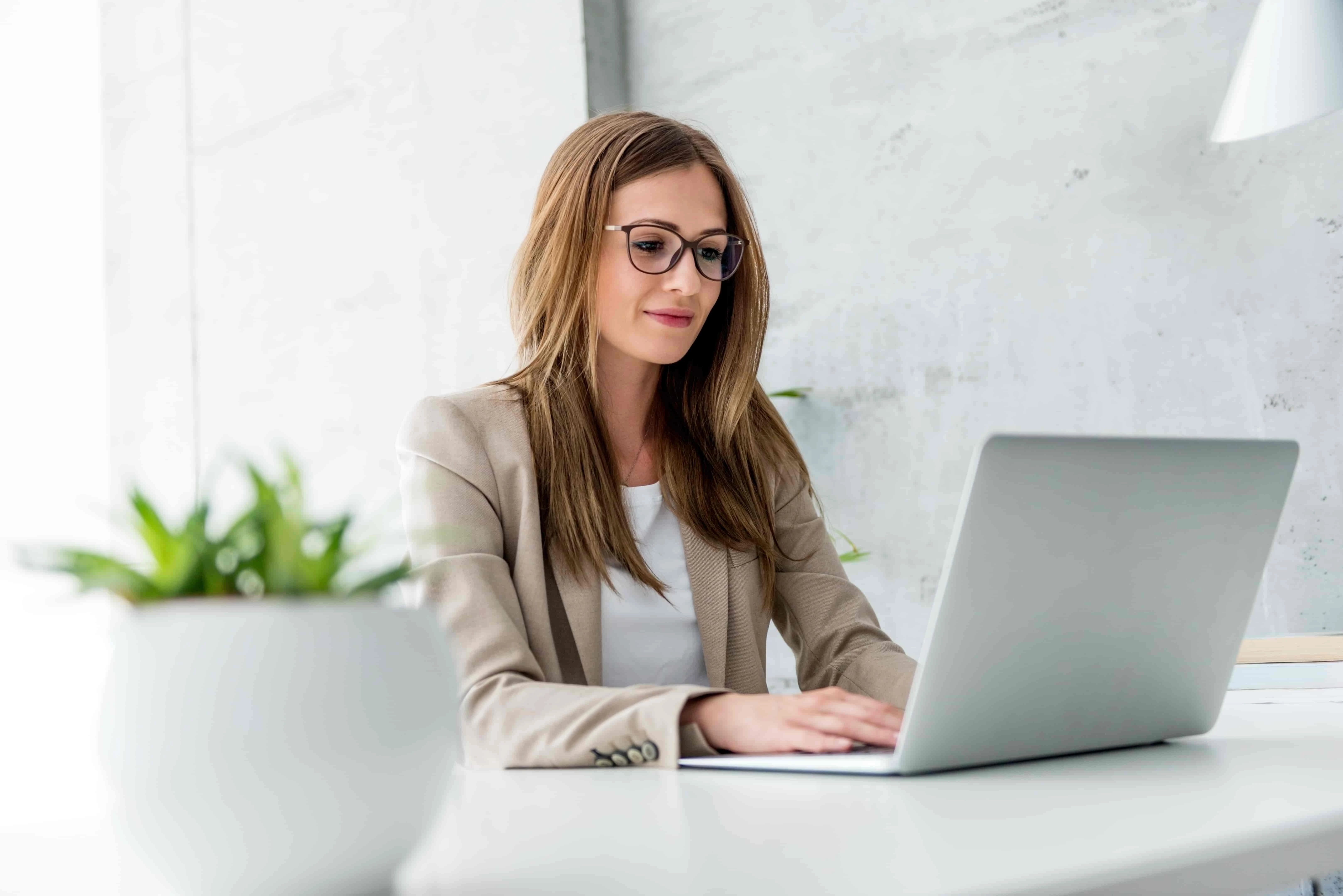 Woman in glasses and a blazer working on a laptop at a modern desk with a plant in the foreground.