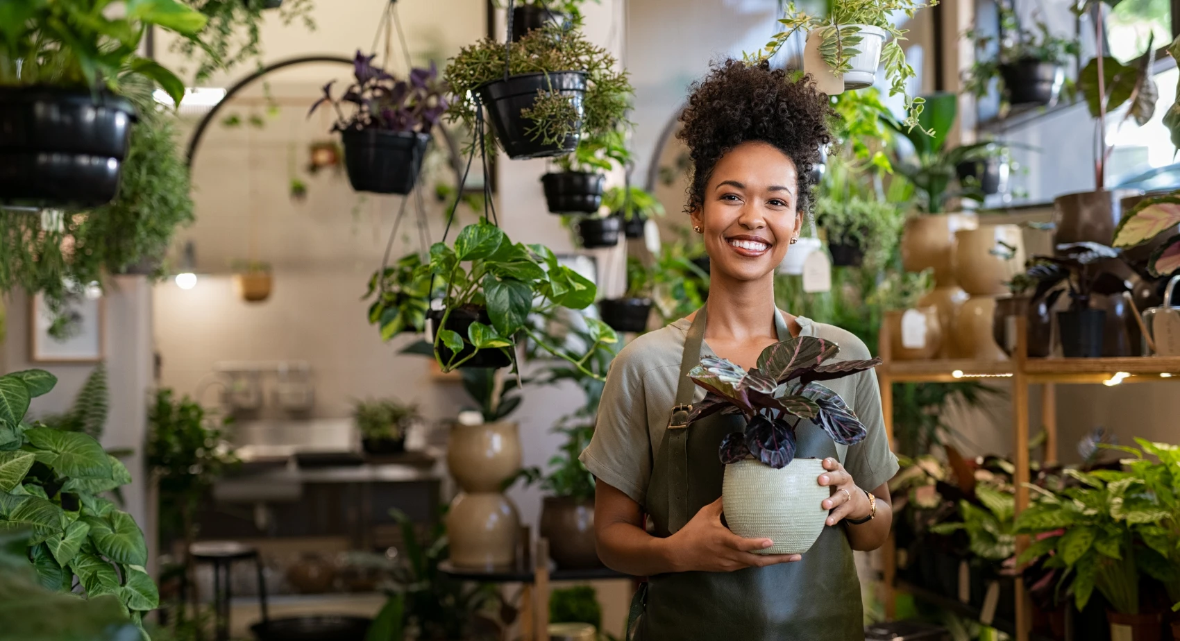 A person smiles warmly, holding a potted plant in a lush, green plant shop. Hanging and potted plants surround them, creating a serene atmosphere.