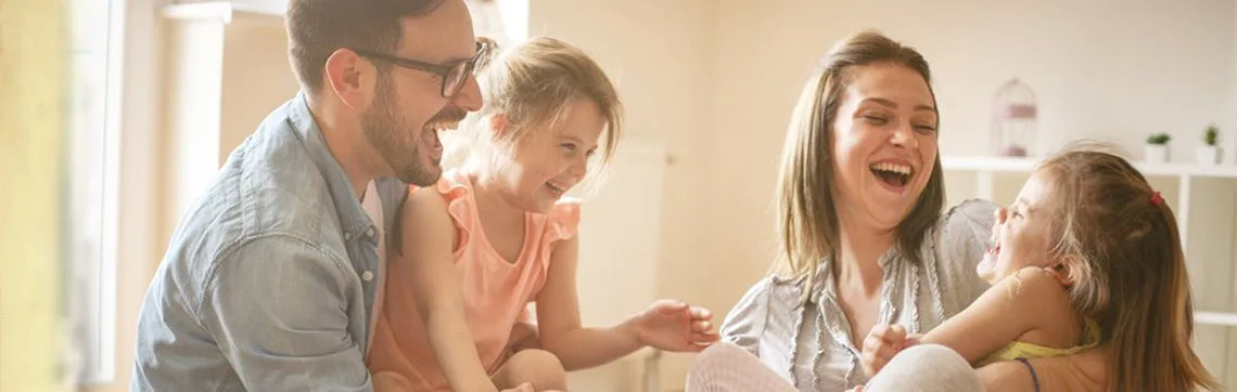 A joyful family of four, including parents and two young girls, are laughing together indoors. The scene conveys warmth, happiness, and connection.