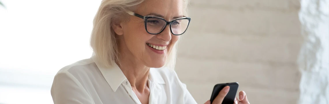 Elderly woman with short white hair and glasses, smiling at her smartphone, wears a white shirt. Background is softly lit with a light brick wall.