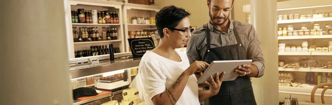 Two small business owners using a tablet while standing in a grocery or deli shop, surrounded by shelves of products.
