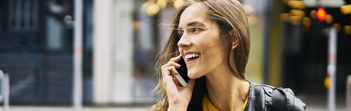 A smiling woman with long hair talks on a smartphone outdoors. She's wearing a leather jacket, conveying happiness. The background is an urban street.