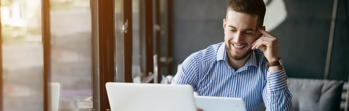 Smiling man in a striped shirt using a tablet and laptop while sitting by a window in a modern office or café.