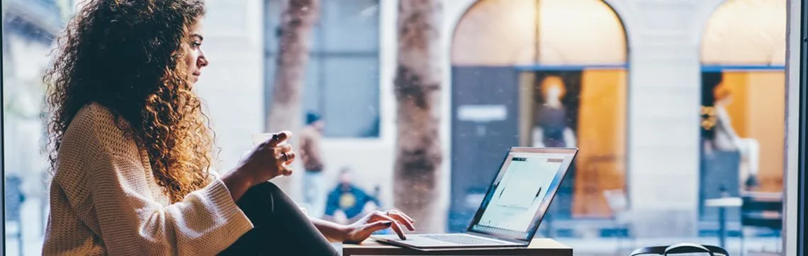 A woman with curly hair sits by a window, holding a mug and using a laptop. She seems focused and relaxed. Outside, blurred buildings create a cozy atmosphere.