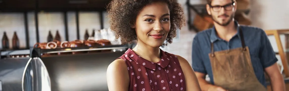 Smiling woman standing confidently in a café or bakery, with a man in an apron in the background.