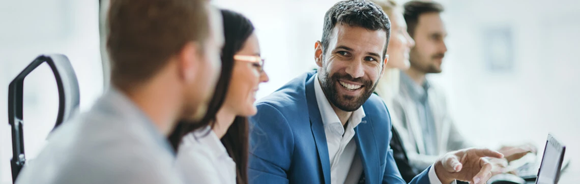 Group of professionals in a meeting, with one man in a blue blazer smiling and engaging in conversation.