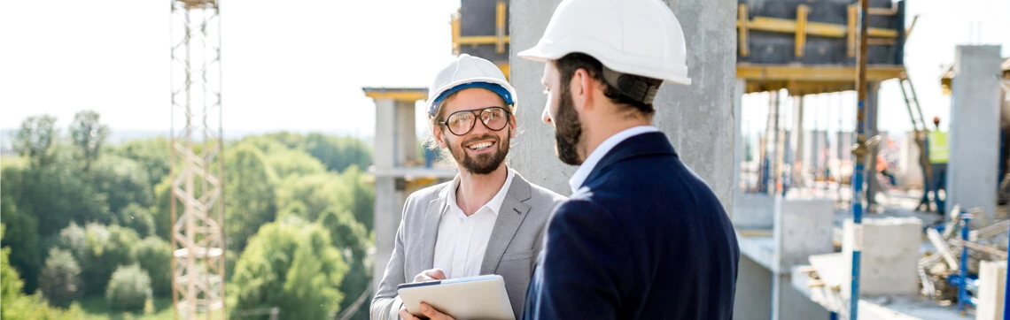 Two men wearing hard hats and suits discussing plans at a construction site, with one holding a tablet.