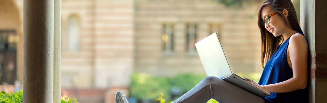 A woman wearing glasses is sitting on a ledge outdoors, using a laptop. She is focused and relaxed, with blurred, sunlit buildings in the background.