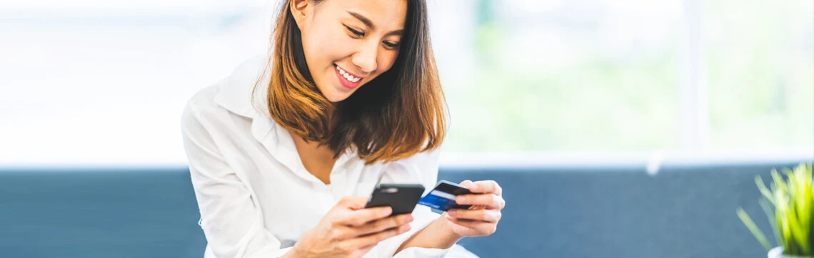 Smiling woman holding a credit card and smartphone while making an online payment.