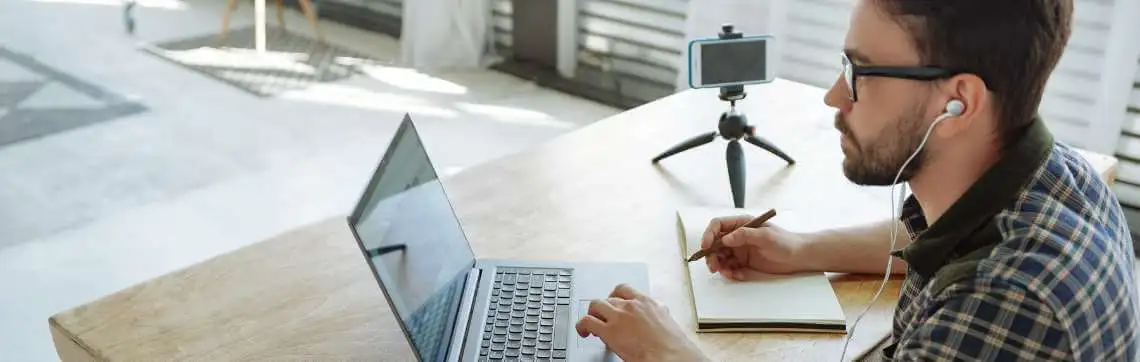 Man with earphones working on a laptop takes notes at a wooden table. Nearby, a smartphone on a tripod suggests a video call setup. Bright, airy room.
