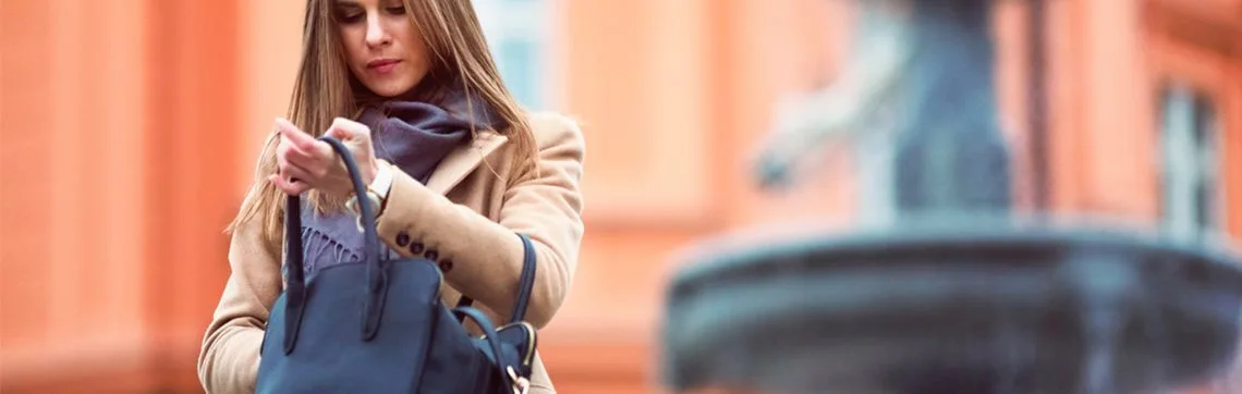 Woman in a tan coat and scarf searches inside a navy handbag. Background features a blurred orange building and fountain, conveying a calm urban scene.