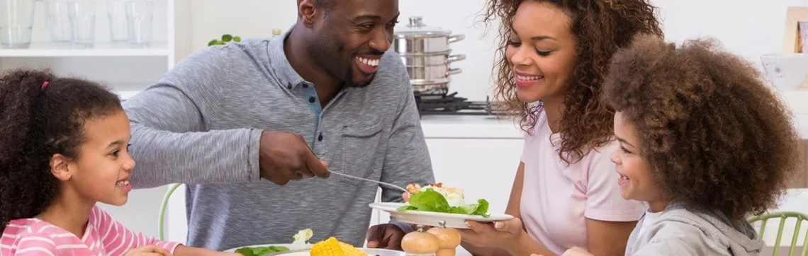 A family of four, smiling and enjoying a meal together at a dining table. Plates have a variety of food, creating a warm and joyful atmosphere.