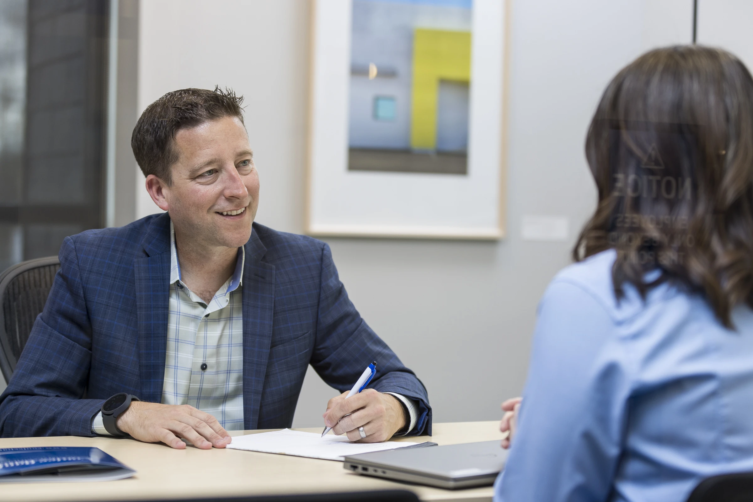 Man in a blue blazer sitting at a desk writing on a document and talking to a woman across the desk who is wearing a blue shirt.