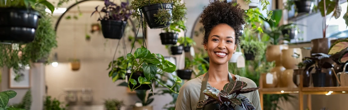 Woman holds a house plant while surrounded by houseplants for sale in her small business.